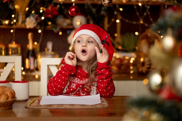 A little cute girl child in a red sweater and a hat in a dark kitchen with a Christmas tree writes a letter to Santa Claus and thinks what to wish for her and waits for the new year or Christmas