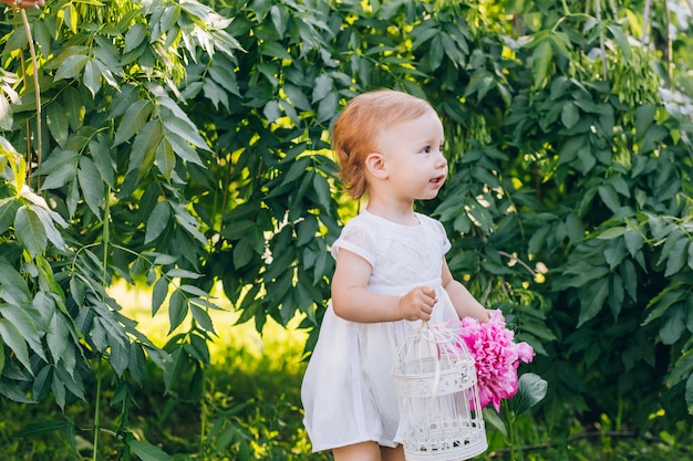 Little cute girl child in a white dress with a pink peony in hands in the park. Portrait of a child outdoors on a sunny summer day. Nice girl holding flowers in her hands.