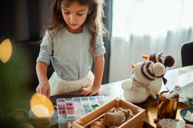 Photo little cute girl chooses buttons for knitted snowmen new years decor is on the table preparation for...