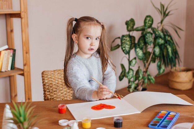 Little cute girl draws with paints at the table in the living room