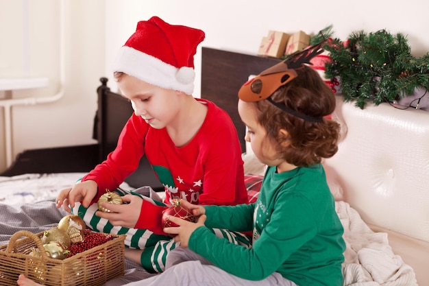 Little cute girl in funny deer mask and boy in Santa cap in Christmas pajamas get toys out of the box and get ready for the holiday.