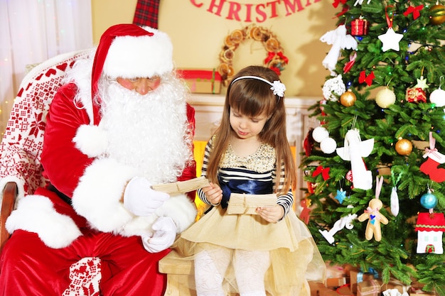 Little cute girl giving letter with wishes to Santa Claus near Christmas tree at home