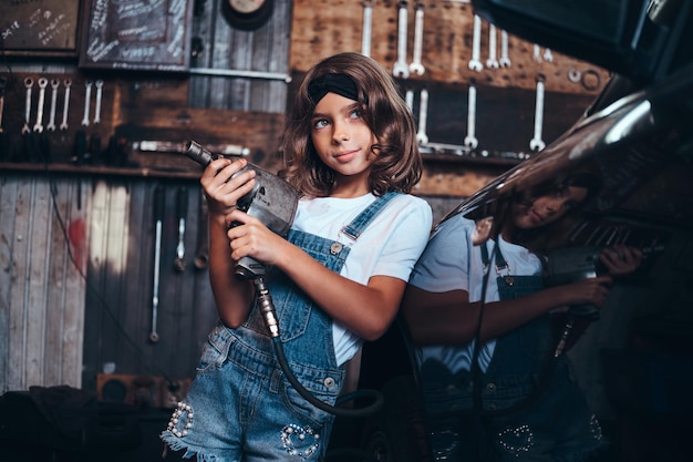 Little cute girl is posing with pneumatic drill for photographer at auto service workshop.