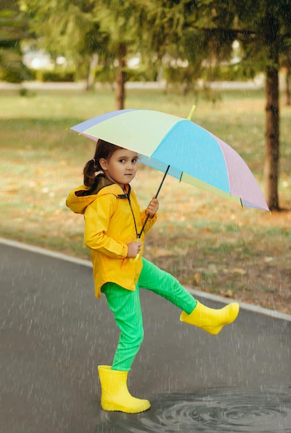 Little cute girl kid child in a yellow raincoat with multicolored rainbow umbrella walking on a puddle