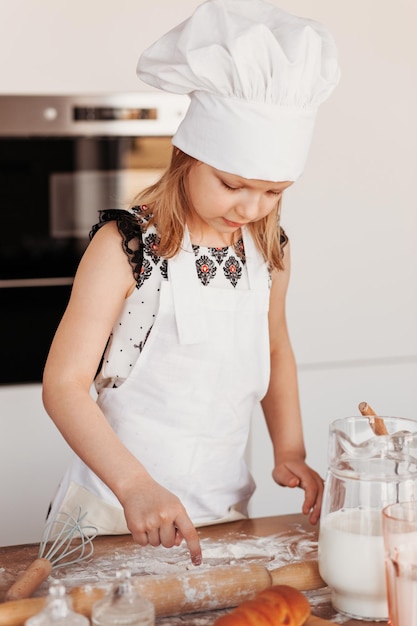 Little cute girl learns to make dough in the kitchen in a white chef's hat