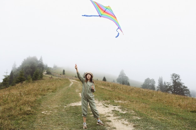 Little cute girl playing with colorful kite on foggy day in the mountains. happy child in green jumpsuit and cap is having fun outdoors