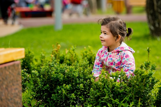 Little cute girl sitting in green bush in the park smiling and looking away