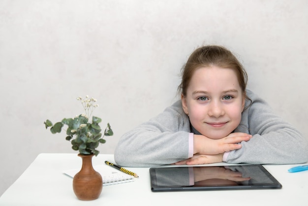 Little cute girl sitting at the table in front of the tablet with her hands folded and smiling learning