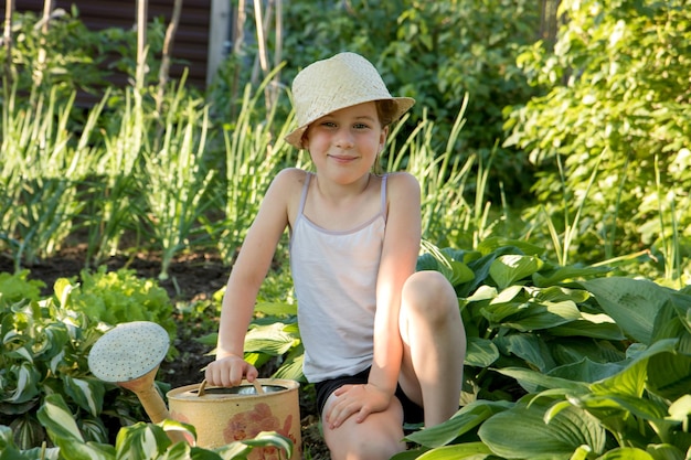 Little cute girl in a straw hat sits next to a watering can surrounded by beds