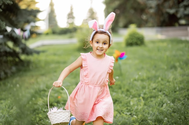 Little cute girl wear bunny ears holding basket with colorful painted eggs on Easter egg hunt in park and running