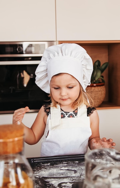 A little cute girl in a white chef hat plays with flour on the kitchen table