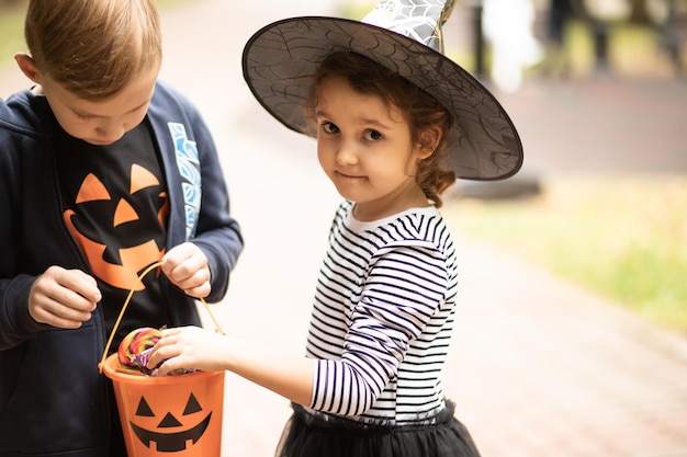 Little cute girl in witch costume and boy holding jack-o-lantern pumpkin bucket with candies and sweets and tastes candy. Kids trick or treating in Halloween holiday.