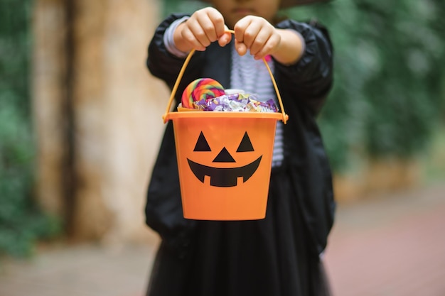 Little cute girl in witch costume holding jackolantern pumpkin bucket with candies and sweets Kid trick or treating in Halloween holiday