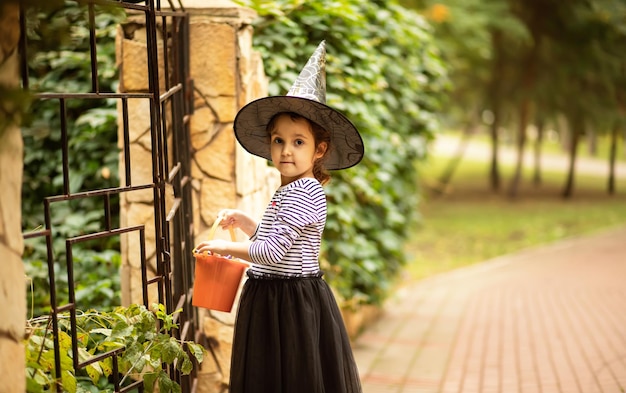 Little cute girl in witch costume holding jackolantern pumpkin bucket with candies and sweets Kid trick or treating in Halloween holiday