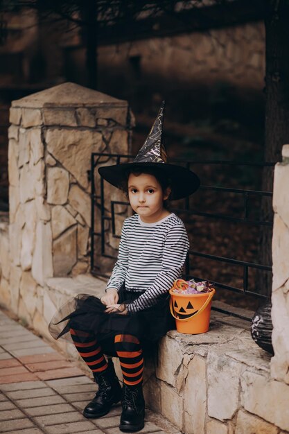 Little cute girl in witch costume holding jackolantern pumpkin bucket with candies and sweets Kid trick or treating in Halloween holiday