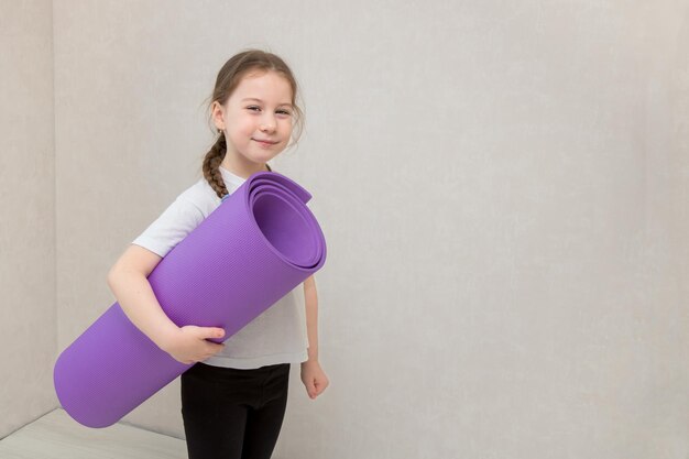 Little cute girl with pigtail holds a gymnastic mat in her hands and smiles with a squint copy space