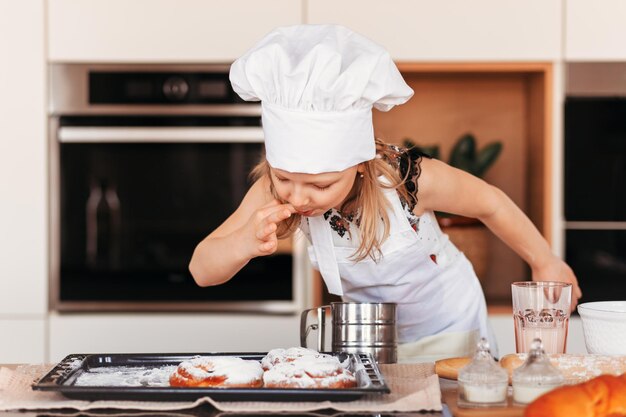 A little cute girl with a white chef hat tastes pastries in the kitchen