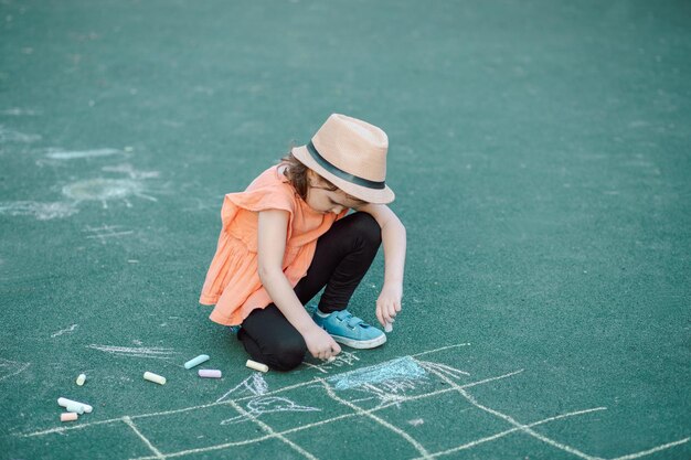 Little cute happy girl in a hat draws painting a chalk on pavement asphalt on ground on playground Creative children summer activity