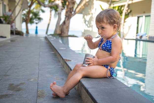 little cute toddler girl in swimsuit eating yogurt sitting by the pool. close up, soft focus