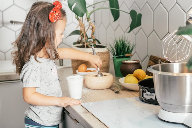 Little dark-haired girl 3 years old in red headband bakes apple pie in kitchen. Child puts sugar in measuring cup