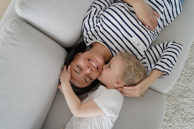 Little daughter kiss smiling mum on cheek relaxing on couch at home Tender moment of motherhood