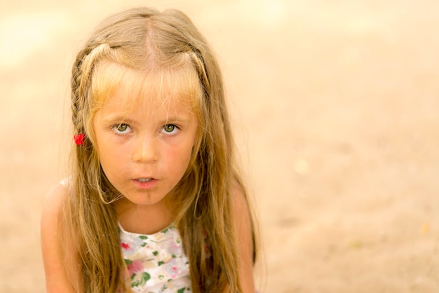 Little dirty blond girl with a pigtail gazes ahead on the background of sand