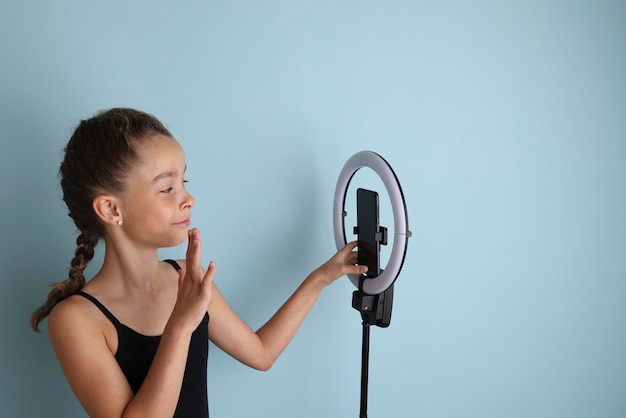 Little emotional teenage girl in a black tshirt on an isolated blue background Studio portrait of a young smiling teenage girl vlogger with a ring flash lamp and a smartphone Girl takes a selfie