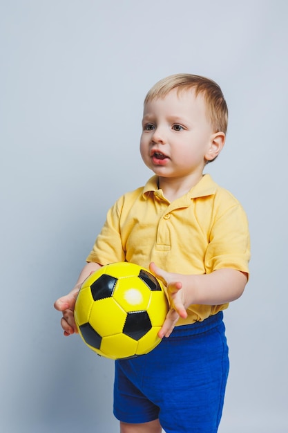 Little european boy fan or player in yellow and blue uniform with a soccer ball supports the soccer team on a white background Football sport game lifestyle concept Isolated on white background