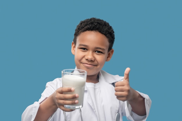 Little expert. Smiling cute darkskinned boy in medical gown with glass of milk showing ok gesture standing on blue background