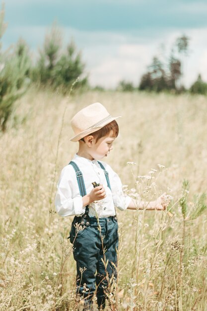 Little farmer in the summer field, cute little boy in a straw hat. boy collects flowers in the field. boy in rubber boots and a white shirt. Country style