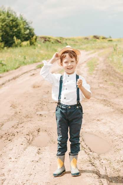 Little farmer on a summer field, cute little boy in a straw hat. boy with a flower stands in a field. boy in rubber boots and a white shirt. Country style