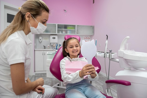 Little female patient in the dental chair looks at her teeth after their treatment dental office mirror in hands