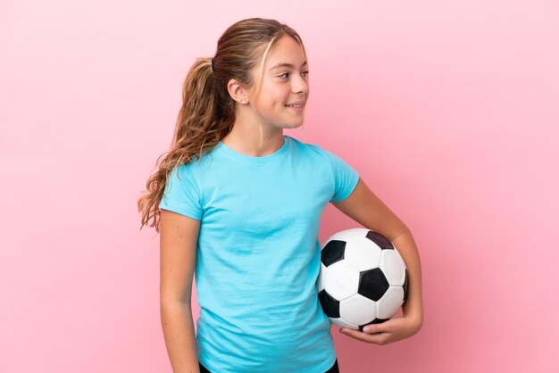 Little football player girl isolated on pink background looking to the side and smiling