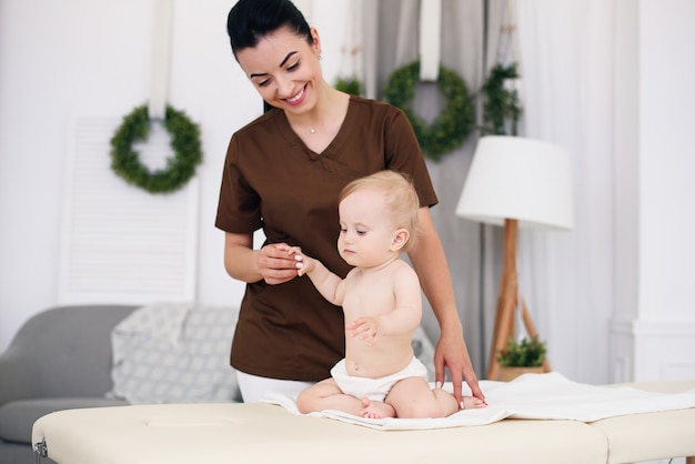 A little funny baby having massage with professional female masseuse. Children's massage on the couch in a modern cozy room. Kind and friendly children's doctor.