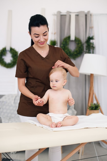 A little funny baby having massage with professional female masseuse. Children's massage on the couch in a modern cozy room. Kind and friendly children's doctor.