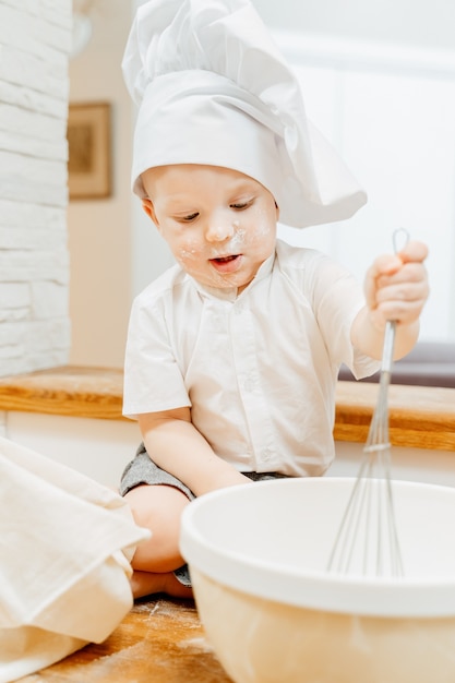 Little funny boy in a cook suit holding whisk with pancake dough