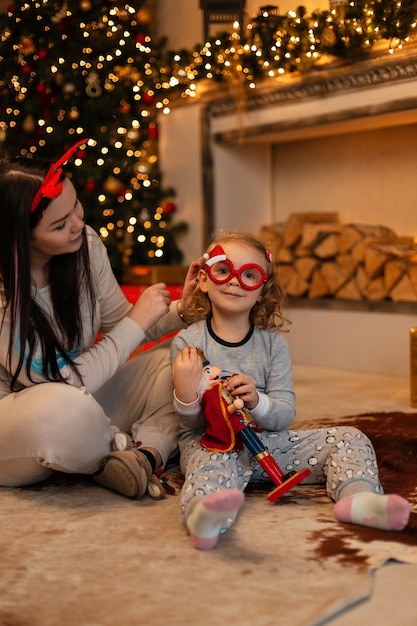 little funny girl with Santa glasses and mom in fashionable festive pajamas sit on the floor near Christmas tree