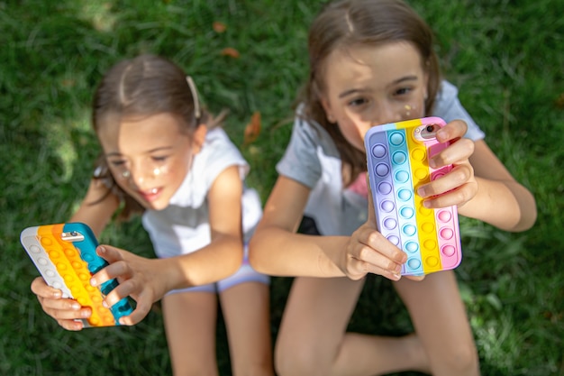 Little funny girls on the grass with phones in a case with pimples pop it, a trendy anti stress toy.