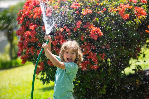 Little gardener child helping to watering flowers with garden hose in summer garden seasonal yard wo