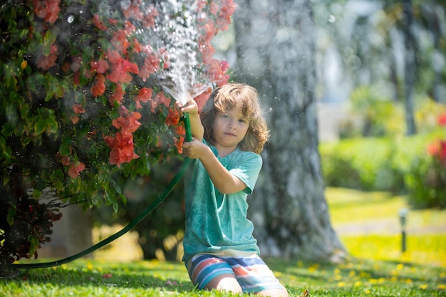 Little gardener child helping to watering flowers with garden hose in summer garden seasonal yard wo