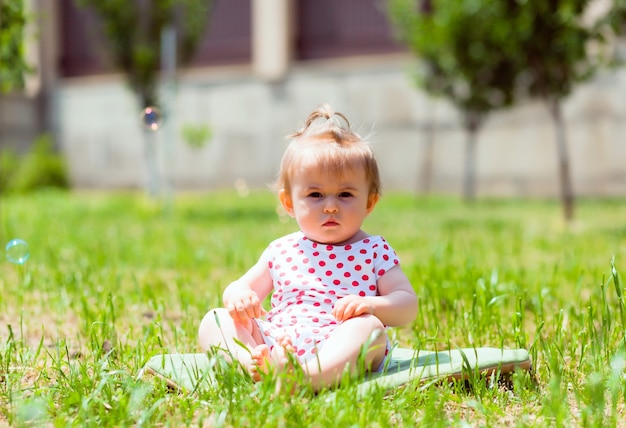 Little girl 11 months old is sitting in the park on the grass. baby girl in a jacket in peas sits on the grass and plays with soap bubbles.