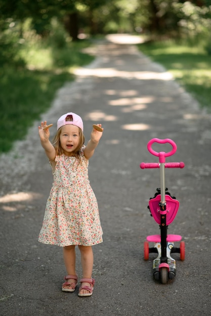 A little girl 3 years old in a pink cap rides a scooter Summer time