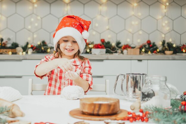 Little girl 3 years old in red Christmas cap and checkered shirt prepares dough for gingerbread cookies in kitchen