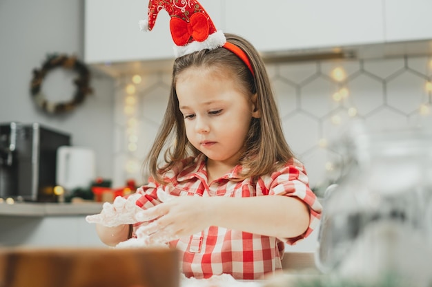 Little girl 3 years old in red Christmas cap and checkered shirt prepares dough for gingerbread cookies in kitchen