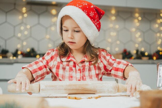 Little girl 3 years old in red Christmas cap and checkered shirt roll out dough for gingerbread cookies in kitchen