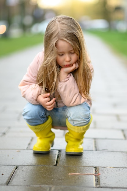 A little girl 3 years old walks in yellow rubber boots after the rain and watches the earthworm