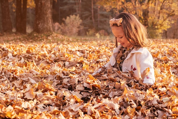 Little girl 34 years old squatting in park in rays of sun and picking up large armful of oak autumn leaves Vertical