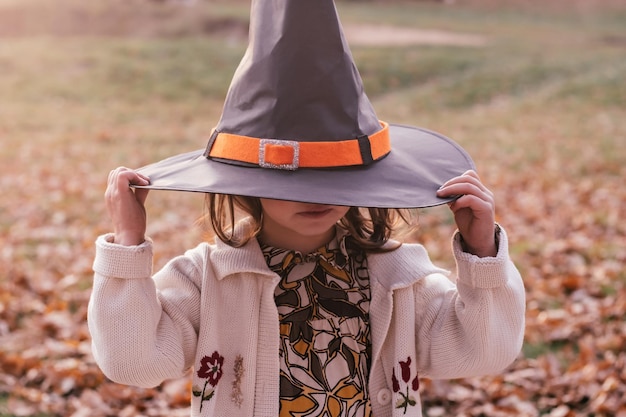 Little girl 34 years old stands outdoor in park on autumn fallen oak leaves and pulls halloween witch hat over her eyes