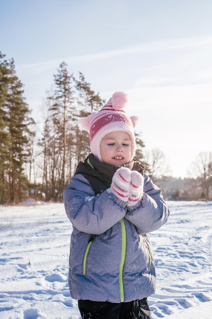 Little girl 34 years old in winter clothes overalls hat and mittens stands outdoors and throws snow laughing Vertical