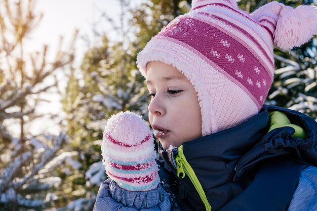 Little girl 34 years old in winter overalls hat and mittens stands against snowcovered pines and fir trees eats snow